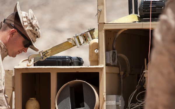 Two military personnel in MARPAT desert camouflage uniforms are examining or working with a modular wooden storage unit containing various equipment. The unit holds compressed gas cylinders, a cylindrical foam-lined container, cables, and a hard-shell case. The setting appears to be an arid outdoor environment, likely a field operation or training exercise.