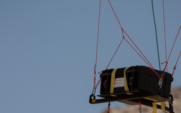 A black hard-shell case or equipment container is suspended in mid-air by a network of red and green rigging lines, secured to a flat platform or pallet with yellow straps. The setup appears to be part of an aerial delivery, parachute drop test, or crane lift operation. A rocky, arid hillside is visible in the blurred background beneath a clear blue sky.