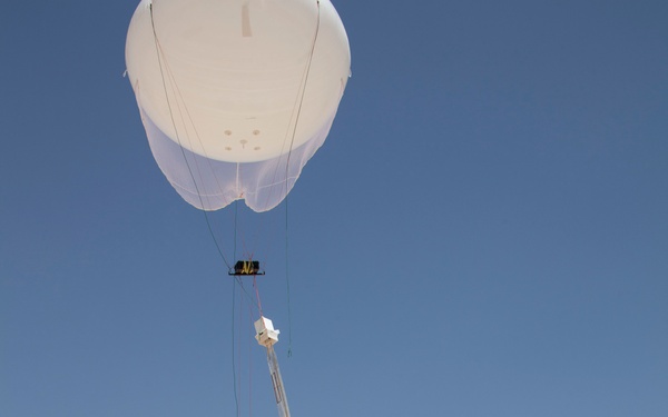 Military personnel in desert camouflage uniforms are launching a tethered weather or surveillance balloon in an arid desert environment. The balloon carries suspended instrumentation packages beneath it, connected by tether lines to a military tactical vehicle on the ground. The scene appears to be a field operation or training exercise in a rocky desert terrain.