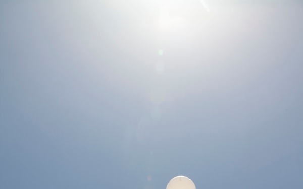 A low-angle upward shot of military personnel in desert camouflage uniforms releasing or observing a white weather balloon ascending into a clear blue sky. The photograph is taken from ground level looking up, creating a dramatic perspective with the sun visible near the top of the frame. The personnel appear to be conducting a meteorological or atmospheric observation activity.