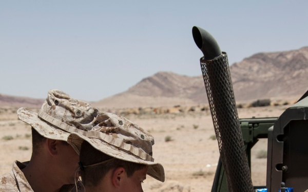 Two military personnel in desert camouflage uniforms (MARPAT desert pattern) are operating a ruggedized Panasonic Toughbook laptop mounted on the rear of a military vehicle, likely a HMMWV (Humvee), in an arid desert environment. The soldiers are wearing boonie hats and appear to be conducting communications or data operations in the field. A radio antenna and handheld radio device are visible alongside the laptop setup.