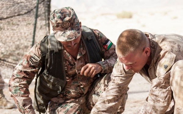 Two military personnel in desert camouflage uniforms are crouched on sandy ground, collaborating on assembling or examining a device mounted on a flat board. One soldier wears a patch identifiable as the Jordanian flag, while the other wears a lighter desert MARPAT-style uniform. Additional military personnel are visible in the blurred background.