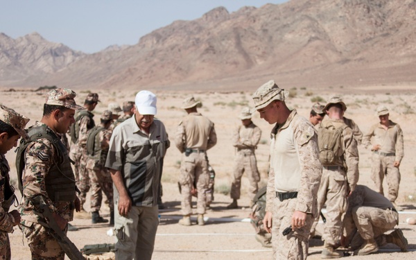 A joint military training exercise taking place in an arid desert environment with mountainous terrain in the background. Personnel in two distinct uniforms — one in multi-terrain camouflage pattern and another in desert MARPAT-style camouflage — are engaged in what appears to be IED detection or demining training using a metal detector probe. A civilian interpreter or advisor in civilian clothing is also present among the group.