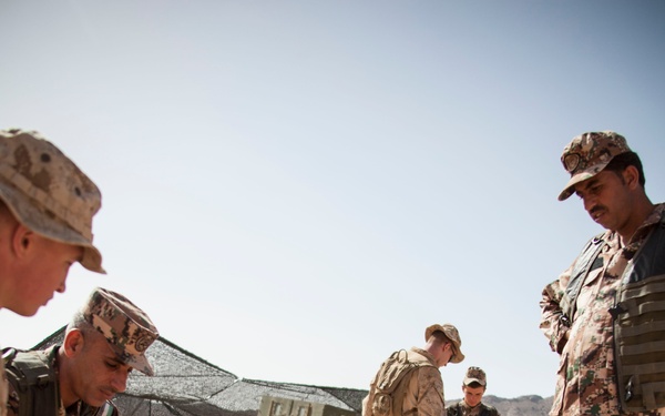 Military personnel from at least two different forces are engaged in what appears to be a mine detection or IED training exercise in a desert environment. One soldier is prone on the ground examining a marked lane with a triangular marker, while others observe and supervise. The scene includes both US Marines (in MARPAT desert camouflage) and soldiers wearing a different digital camouflage pattern with what appear to be Jordanian Armed Forces insignia.