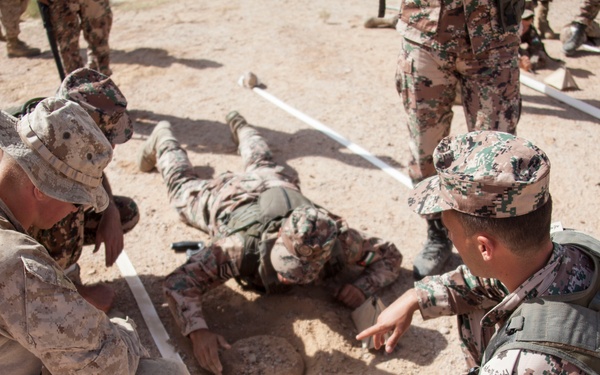 Military personnel from two different forces are engaged in a joint training exercise on sandy terrain. One soldier is lying prone on the ground while others crouch nearby, appearing to examine or demonstrate something related to ground-level detection or IED awareness training. White tape lines are visible on the ground, likely marking a training lane or boundary.