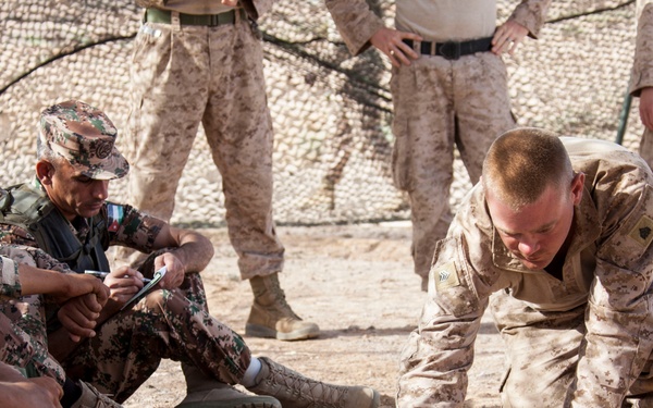 A military training or briefing session is taking place in an outdoor desert environment. A soldier in MARPAT desert camouflage is kneeling on the ground, appearing to demonstrate or explain something using the dirt surface, while other soldiers in mixed camouflage uniforms observe. The setting suggests a joint multinational military exercise or training event.