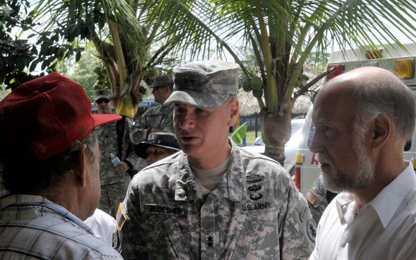 A U.S. Army soldier, identified by name tape as 'RODESHEIM' and wearing ACU (Army Combat Uniform), is engaged in conversation with a civilian man in a plaid shirt and red cap, alongside an older civilian man in a white shirt. The setting appears to be an outdoor location in a Spanish-speaking country, evidenced by a vehicle in the background marked 'CENTRO DE SALUD TORTI' (Torti Health Center). Multiple other military personnel are visible in the background.