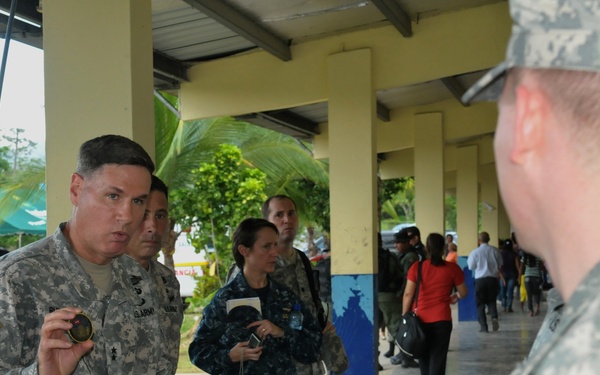 A group of U.S. military personnel in ACU (Army Combat Uniform) and Navy working uniforms are gathered under a covered outdoor corridor of what appears to be a medical or civic facility in a Spanish-speaking country. A senior Army officer, identifiable by rank insignia, is holding up what appears to be a challenge coin while speaking to another soldier whose name tape reads 'ERWIN'. The setting suggests a humanitarian mission or medical readiness exercise, supported by a visible 'Salud' (health) sign in the background.