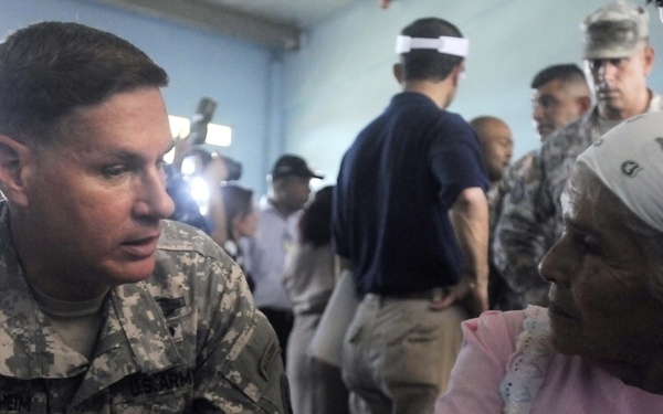 A U.S. Army soldier, identified by name tape as 'Rudenheim,' is seated at a table speaking with an elderly woman wearing a patterned headscarf and pink clothing. Several medication bottles are visible on the table, suggesting a medical assistance or humanitarian aid context. Additional military personnel and civilians are visible in the background of what appears to be an indoor facility.