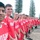 A large group of young male soccer players wearing matching red 'Hooligans' jerseys by Vizari are lined up in a row for a team photo. The photo appears to be taken as a wide-angle selfie by the player on the far left, creating a perspective distortion along the line. The setting appears to be an outdoor sports facility with tall Norfolk pine trees visible in the background.