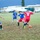 A recreational soccer match is being played on a grass field between two teams — one in blue uniforms labeled 'HOOLIGANS' and one in red uniforms. Players are actively competing for the ball near the center of the field, with visible jersey names including 'REEVES #44' and 'SANCHEZ #18'. The setting appears to be a military or institutional facility based on the buildings and vehicles visible in the background.
