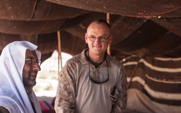 The image shows a meeting or conversation taking place inside a traditional Bedouin-style tent. A man wearing a white keffiyeh and dark robe is engaged in discussion with individuals in military camouflage uniforms. The setting suggests a civil-military engagement or liaison meeting in a desert region.