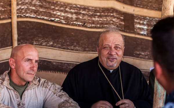 Three individuals are seated together inside what appears to be a traditional Bedouin-style tent with a striped fabric backdrop. Two figures face the camera: one wearing US Marine Corps MARPAT desert camouflage uniform and one wearing black clerical robes with a gold cross pectoral, suggesting a Christian Orthodox or Eastern Catholic clergy member. A third person in blue digital camouflage (likely US Navy NWU) is visible from behind in the foreground.