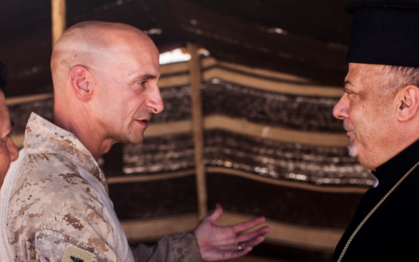 A military officer in desert camouflage uniform is shaking hands with a religious cleric wearing black robes and a black clerical hat with a gold chain/cross. A third individual in a dark naval uniform is visible on the left side of the frame. The setting appears to be inside a tent or temporary structure.