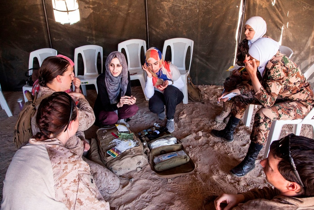 A group of women are gathered in a circle inside a military-style tent, appearing to be engaged in a discussion or briefing session. The group includes women in Western military uniforms (desert camouflage pattern consistent with US Marines) and women in civilian clothing wearing hijabs, as well as at least one woman in what appears to be Jordanian military camouflage. Medical or tactical gear bags are visible on the sandy ground in the center of the group.