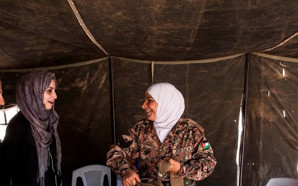 The image shows a group of four women inside a military tent, appearing to be engaged in a friendly interaction. Two women are wearing Jordanian Armed Forces camouflage uniforms with visible 'JORDAN' name tapes and Jordanian flag patches, while one woman wears what appears to be a Canadian Armed Forces uniform, and another is in civilian clothing. The atmosphere appears relaxed and cooperative, suggesting a joint military or humanitarian engagement.