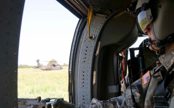 A U.S. Army soldier in full combat gear and flight helmet is seated inside a military helicopter, manning what appears to be a door-mounted weapon system. The soldier is wearing ACU (Army Combat Uniform) pattern camouflage with a visible U.S. flag patch and a red cross patch, indicating a possible medevac role. In the background through the open door, another military helicopter (likely a UH-60 Black Hawk) is visible on a grassy field.