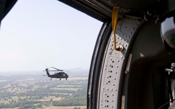A U.S. Army soldier in full flight gear and ACU camouflage uniform is positioned at the open door of a military helicopter, with a mounted machine gun visible in the foreground. Through the open door, a second UH-60 Black Hawk helicopter is visible flying in formation over a rural, flat landscape. The image appears to be taken during a training or operational flight mission.