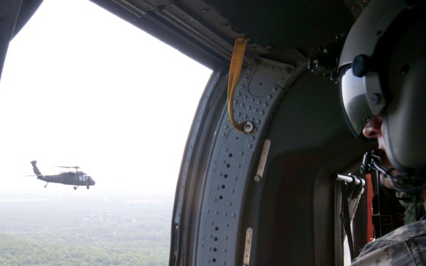 A military door gunner in digital camouflage uniform and flight helmet is positioned at the open door of a helicopter, manning a mounted machine gun. A second helicopter, identifiable as a UH-60 Black Hawk, is visible flying in formation through the open door. The scene appears to be a military training or operational flight over a forested area.