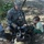 A U.S. Army soldier named Shepherd, wearing ACU (Army Combat Uniform) with an American flag patch, crouches beside a young toddler outdoors. The soldier is showing the child a mounted military weapon system, likely an M240 machine gun on a tripod mount. The interaction appears to be at a public military demonstration or family day event.