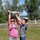 Two women are working together outdoors on a grassy field, jointly lifting a military-style sandbag overhead. One woman wears a pink tank top with military camouflage pants and tactical boots, while the other wears a gray t-shirt and jeans. Both are wearing hydration backpacks, and additional sandbags are visible on the ground nearby.