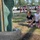 Two young girls with face paint are playing outdoors near a repurposed tire and green metal sculpture at what appears to be a community event or fair. One girl is crouching and holding a water balloon while the other stands beside her, both appearing engaged in a water balloon activity. A parking lot with multiple SUVs and trucks is visible in the background.