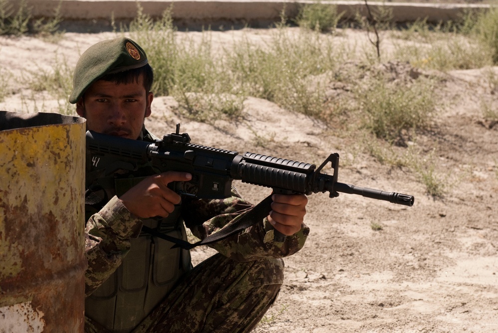 A soldier in camouflage uniform and green beret is positioned behind cover, aiming an assault rifle in what appears to be a training exercise. The soldier is crouched beside a yellow and rust-colored barrier in an arid, sandy environment with sparse vegetation. The image captures a tactical shooting stance with the weapon raised to eye level.