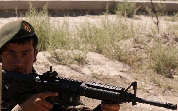 A soldier in camouflage uniform and green beret is positioned behind cover, aiming an assault rifle in what appears to be a training exercise. The soldier is crouched beside a yellow and rust-colored barrier in an arid, sandy environment with sparse vegetation. The image captures a tactical shooting stance with the weapon raised to eye level.