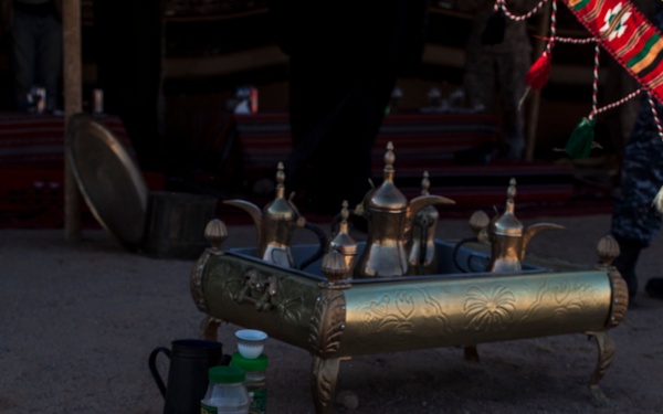 A traditional Arabian desert camp coffee preparation scene showing a metal kettle heating over an open fire made with rocks and embers. In the background, multiple ornate brass dallah (Arabic coffee pots) are displayed on an engraved brass tray with legs. The setting appears to be a Bedouin-style hospitality area with traditional textiles visible in the background.