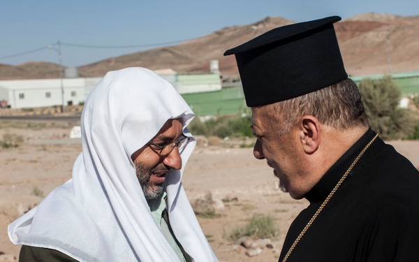 Two religious figures are engaged in a close, respectful greeting or conversation in an arid, desert-like outdoor setting. One individual wears Islamic clerical attire including a white keffiyeh and olive robe, while the other wears Eastern Orthodox or Eastern Catholic clerical vestments including a black cassock, pectoral cross, and kalimavkion (black cylindrical hat). A third person in camouflage military attire is partially visible in the background.