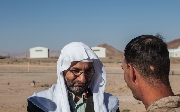 Two individuals are engaged in a handshake in an arid, desert environment. One person is wearing traditional Islamic clerical attire including a white keffiyeh and dark robes, while the other is wearing desert camouflage military uniform with visible sergeant rank insignia. The setting appears to be a remote desert location with small structures and a building with a green dome visible in the background.