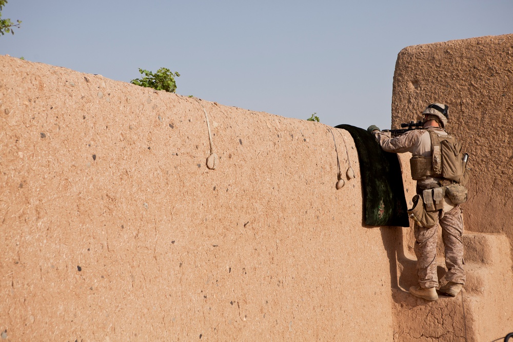 A soldier in desert camouflage uniform is positioned at the top of a mud/adobe wall, aiming a rifle over the wall while standing on steps. The setting appears to be an arid environment with a clear sky, consistent with a Middle Eastern or Central Asian operational theater. The soldier is equipped with full combat gear including body armor, helmet, and tactical pouches.