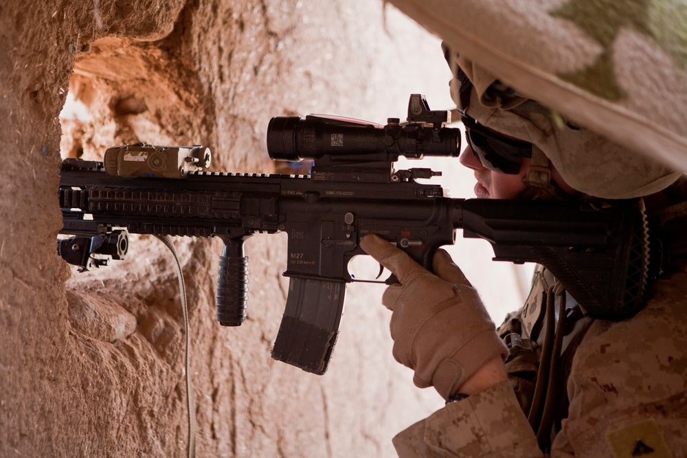 A military service member in desert camouflage uniform is positioned against a mud/adobe wall, aiming a rifle through an opening or loophole. The weapon is clearly marked 'M27' on the lower receiver, identifying it as an M27 Infantry Automatic Rifle (IAR). The soldier is equipped with tactical optics, accessories, and protective eyewear consistent with U.S. Marine Corps combat gear.