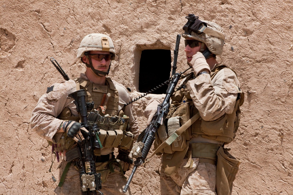 Two military personnel in desert combat uniforms are positioned against a mud/adobe wall in what appears to be an arid environment. Both are fully equipped with tactical gear, body armor, and assault rifles. One soldier is using a radio handset while the other maintains a security posture.