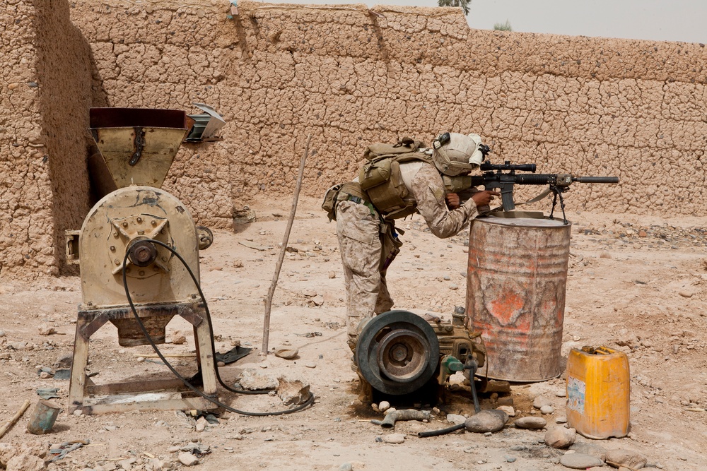 A soldier in desert camouflage uniform is using a rusted metal barrel as a rest while aiming a scoped rifle. The scene is set in an arid environment with a mud-brick wall in the background. Various pieces of equipment and machinery are visible in the compound area.