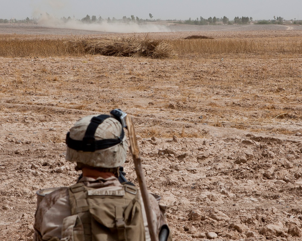 A military soldier in desert camouflage uniform is observed from behind, surveying an arid, open landscape. The soldier is equipped with a rifle and full combat gear, facing toward a dusty field with sparse dry vegetation and trees visible in the distance. A dust cloud is visible on the horizon, suggesting vehicle or activity movement in the background.