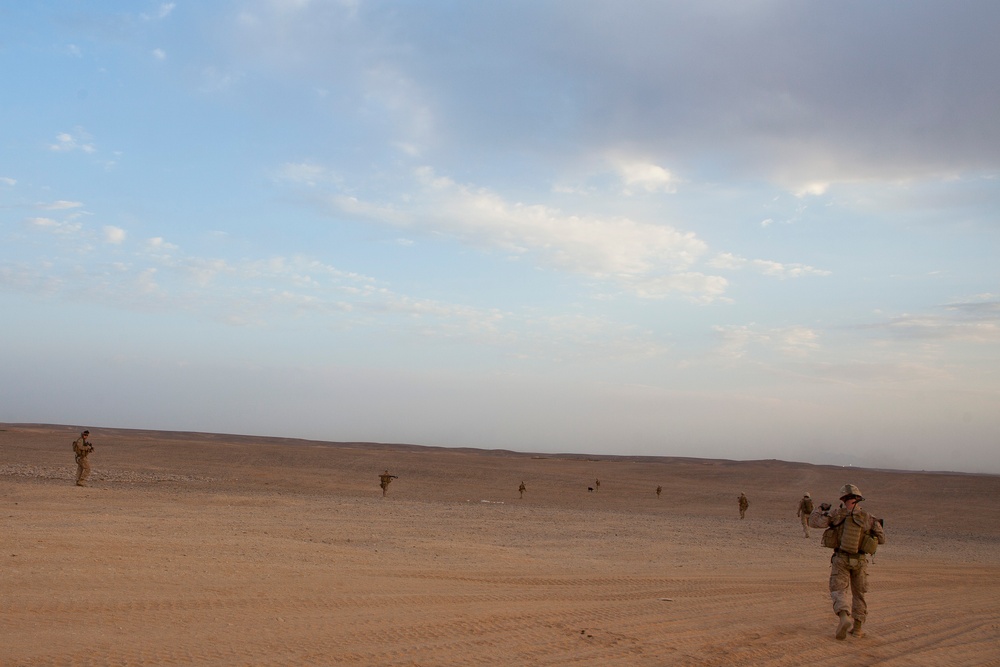 A group of military personnel in desert camouflage uniforms are dispersed across a flat, arid desert landscape. The soldiers appear to be conducting a patrol or tactical movement in a spread formation across open terrain. The scene is captured during daylight with partly cloudy skies overhead.