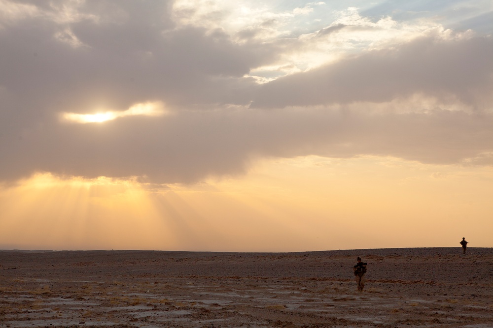 A vast, flat desert landscape is captured during sunset or sunrise, with dramatic golden light rays breaking through heavy cloud cover. Two human silhouettes are visible in the middle distance — one appears to be carrying a large pack or equipment, and another stands further in the background. The scene conveys isolation and scale, with the expansive sky dominating roughly two-thirds of the frame.
