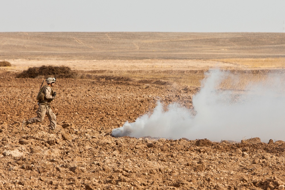 A single soldier in desert camouflage uniform is observed walking or moving across a rocky, arid terrain. White smoke is rising from the ground nearby, consistent with a smoke grenade or controlled detonation. The landscape is flat and barren, characteristic of a desert or semi-arid environment.