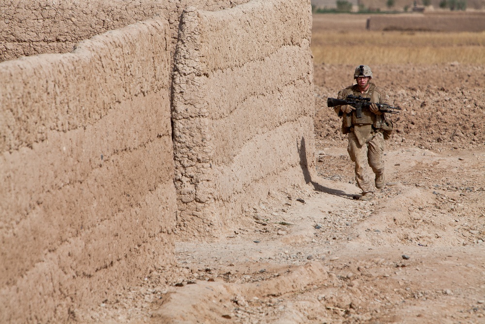A uniformed soldier carrying a rifle moves along a narrow dirt path between mud-brick compound walls in an arid rural environment. The terrain and architecture are consistent with a combat deployment zone, likely in Central or South Asia. Agricultural fields are visible in the background, suggesting proximity to a rural settlement.