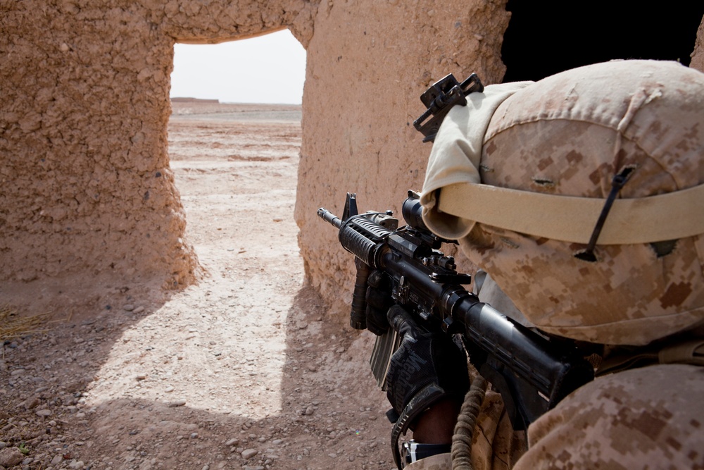 A military service member in desert camouflage (MARPAT pattern) is positioned against a mud-brick wall, aiming a rifle through a doorway opening. The setting appears to be an arid, desert environment with adobe or mud construction typical of Middle Eastern or Central Asian regions. The soldier is in a tactical clearing or security posture near a structure entrance.