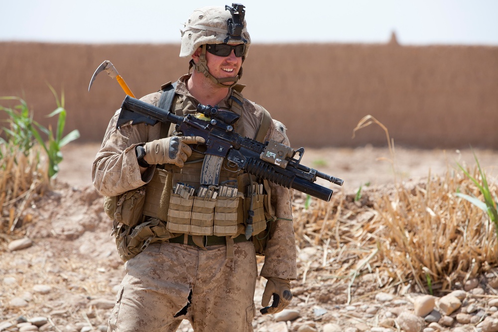 A military service member in desert camouflage uniform is walking through an arid, rural environment. The soldier is fully equipped with body armor, tactical vest with magazine pouches, helmet, and is carrying an assault rifle with an underbarrel attachment. The setting appears to be a dry, dusty area with sparse vegetation and a mud-brick wall visible in the background.