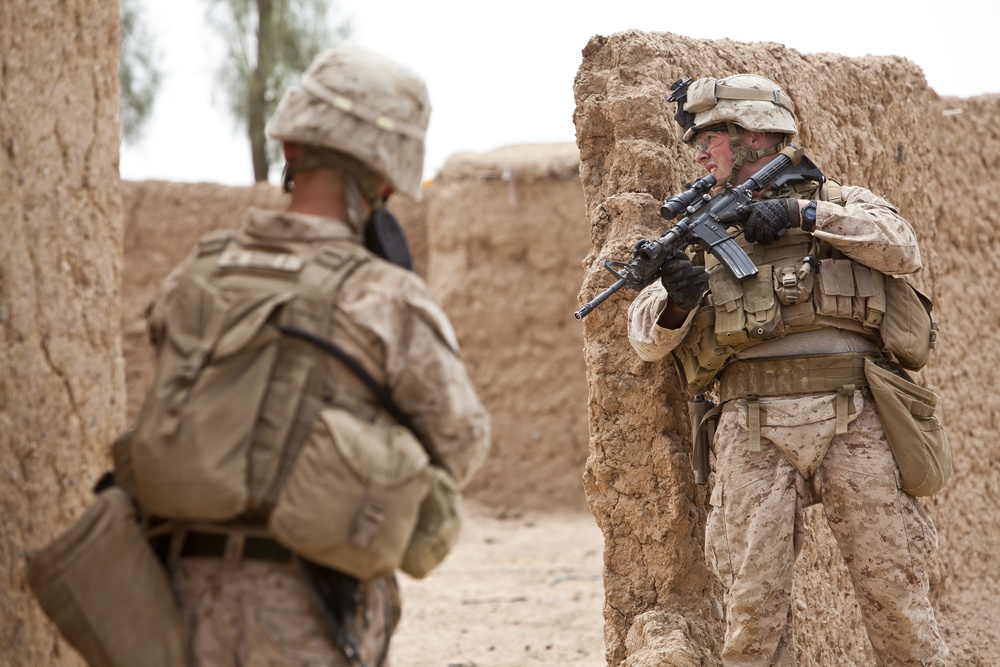 Two military personnel in desert camouflage uniforms are conducting what appears to be a tactical operation in an arid, mud-brick compound environment. The soldier in the foreground right is taking a covered position at a wall corner while aiming a rifle with an optical scope. The setting and uniforms are consistent with operations in a desert region.
