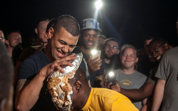 A group of people gathered at night are participating in what appears to be a pie-in-the-face activity or food challenge. One person in a yellow shirt is having a pie or food item pressed into their face by another individual, while a crowd watches and records with smartphones. The atmosphere appears festive and celebratory.
