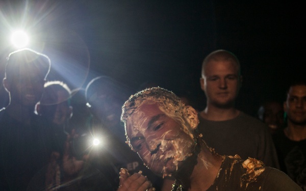 A nighttime outdoor scene showing a man with his face and shoulders covered in what appears to be cake or food substance, surrounded by a crowd of onlookers. Bright camera flashes and lights illuminate the scene against a dark background. Someone is offering a white cloth or towel to the man, likely to help clean up.