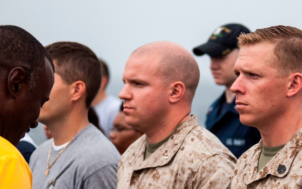 The image shows a group of military personnel and civilians gathered outdoors, likely during a formal ceremony or formation. Two U.S. Marines in desert MARPAT camouflage uniforms are prominently visible in the foreground, with one holding a blue folder containing documents. Additional personnel in Navy uniforms and civilian attire are visible in the background.