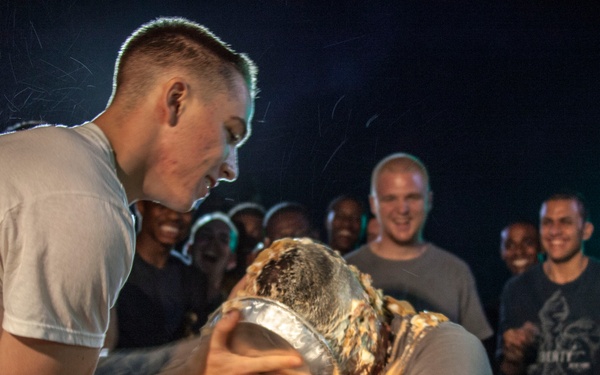 A group of people are gathered outdoors at night watching a celebratory event where one person is having what appears to be a pie or food item smashed into their face. The crowd in the background is laughing and smiling, suggesting this is a lighthearted, consensual activity. The scene appears to be a birthday or initiation-style celebration.