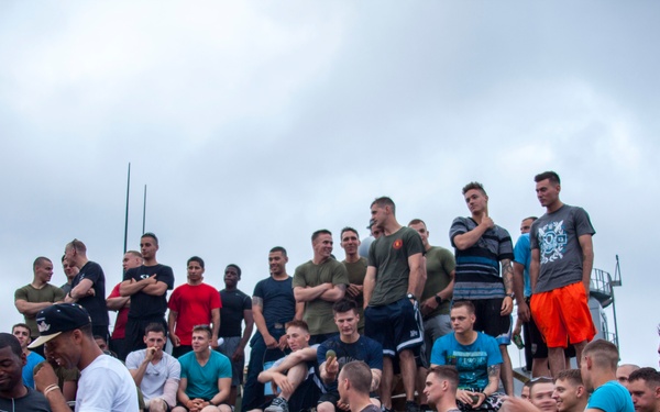 A large group of military personnel, dressed in casual athletic and civilian clothing, are gathered on what appears to be the deck of a naval vessel. Some individuals are standing on top of a military vehicle (likely an armored personnel carrier or similar equipment) chained to the deck, while others stand and sit around it. The overcast sky and ship deck setting suggest an informal gathering or recreational event aboard a ship.