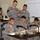 A group of U.S. Army soldiers are engaged in a classroom training session. Several soldiers are seated at desks working on documents while two standing soldiers, appearing to be instructors or NCOs, oversee and instruct the group. A whiteboard with numerical data is visible in the background, suggesting a fire mission or navigation training exercise.