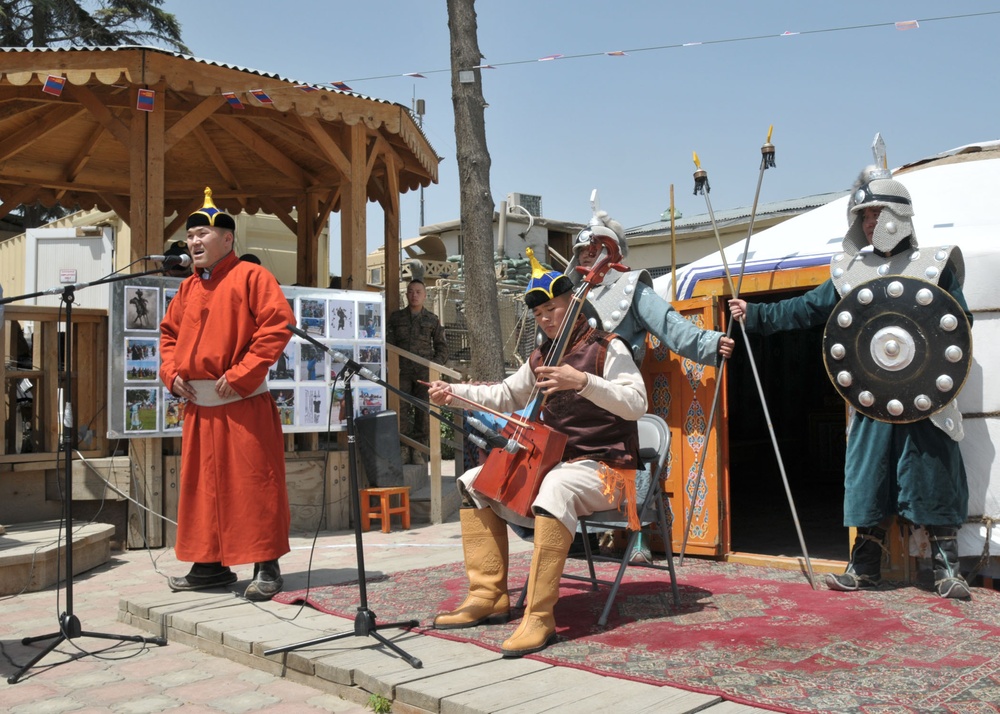 A traditional Mongolian cultural performance is taking place outdoors, likely at a military base or international event given the presence of soldiers in the background. Performers are dressed in traditional Mongolian attire including a singer in a red deel robe, a musician playing a morin khuur (horsehead fiddle), and two guards in historical Mongolian warrior armor holding spears and shields. A traditional ger (yurt) is visible in the background alongside a wooden gazebo.